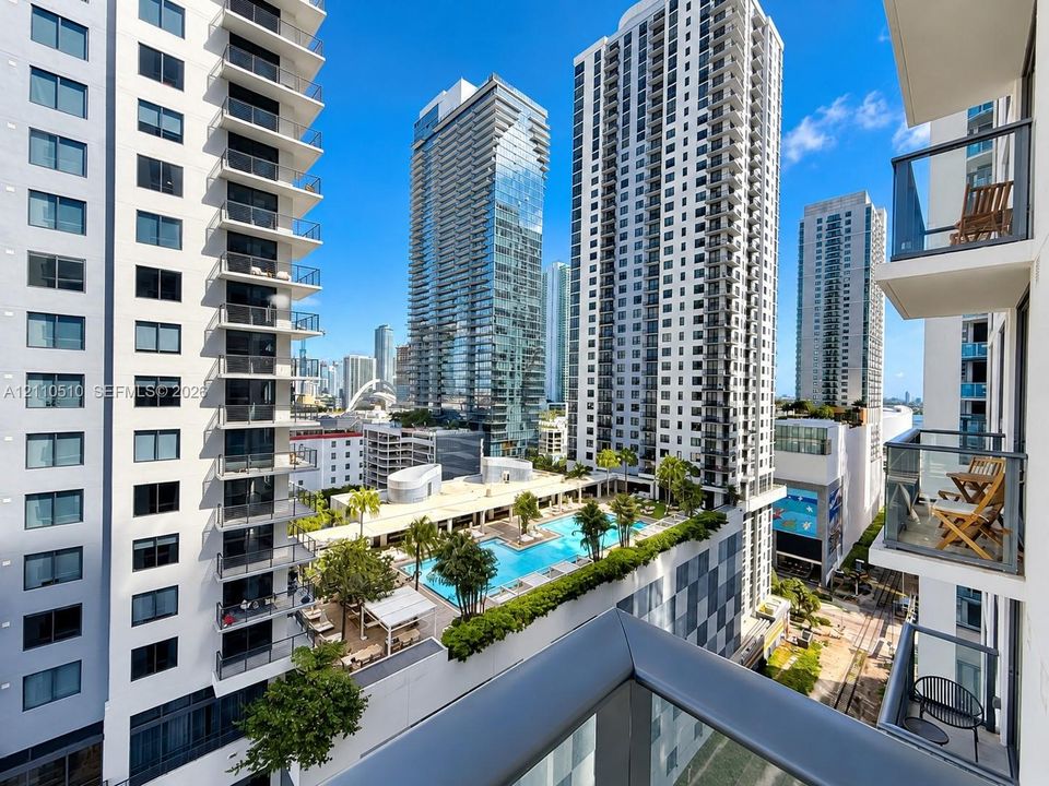 Private balcony with glass panel railing overlooking an outdoor elevated swimming pool, surrounded by high-rise urban architecture