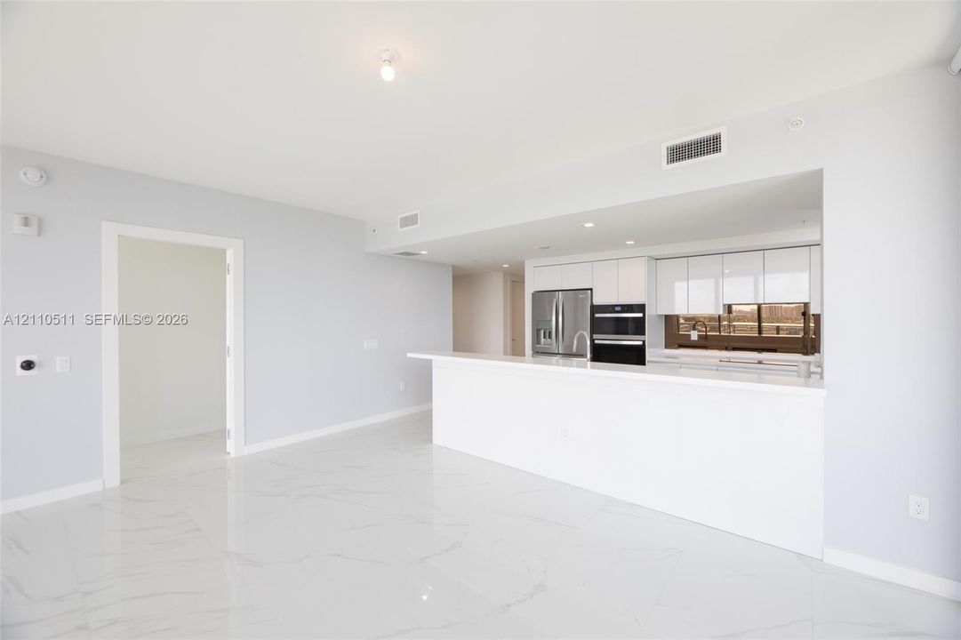 Open-concept area featuring white tile flooring with veining, white flat-panel cabinetry, a stone-surface peninsula, stainless steel appliances, and integrated recessed lighting