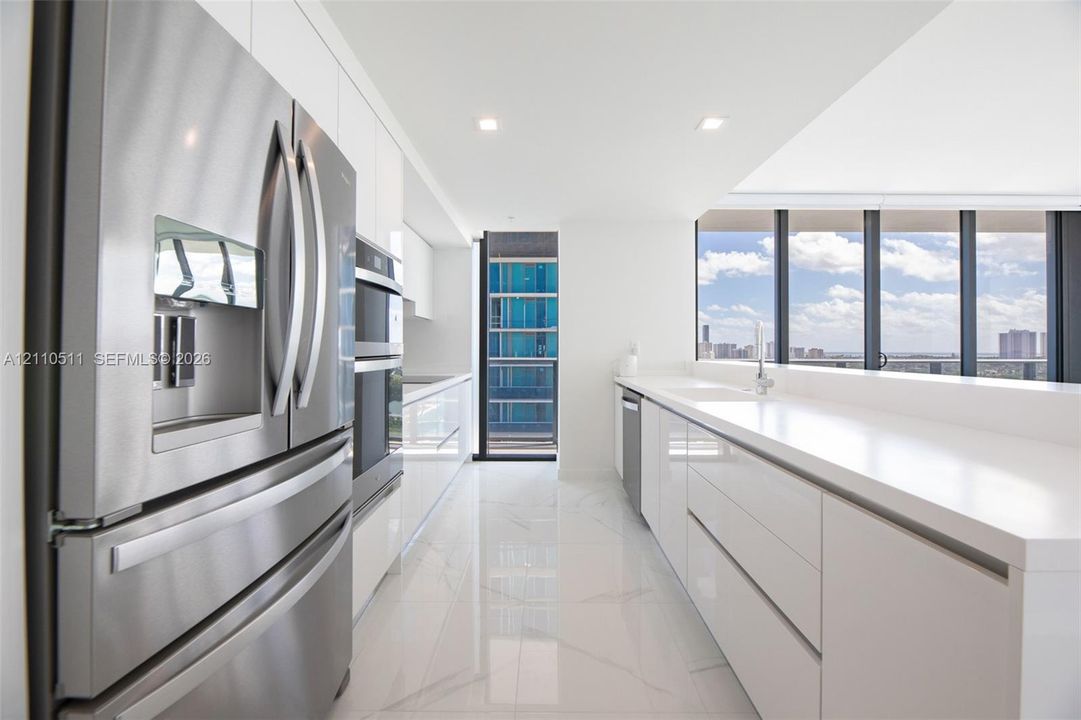 Modern kitchen featuring high-gloss white cabinetry, expansive stone countertops, stainless steel appliances, floor-to-ceiling windows, and large-format tile flooring