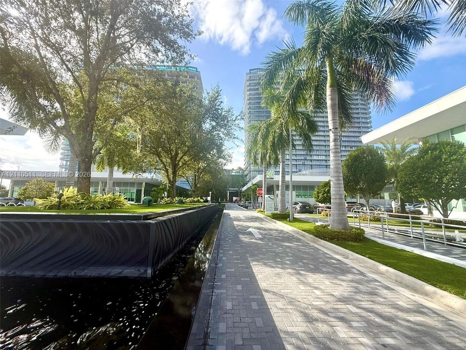 Paver walkway flanked by a water feature with textured cladding, palm tree-lined landscaping, and manicured green space