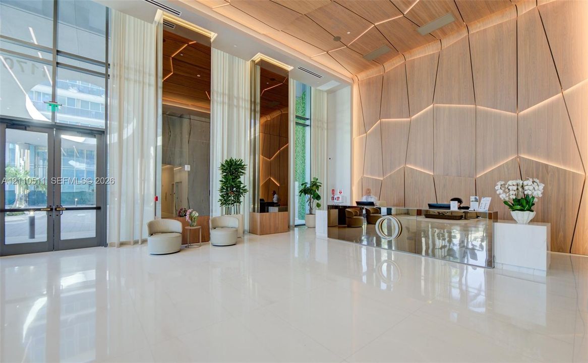Grand lobby featuring polished white tile flooring, floor-to-ceiling glass entryway doors, geometric wood-paneled walls with integrated recessed lighting, and a mirrored reception desk
