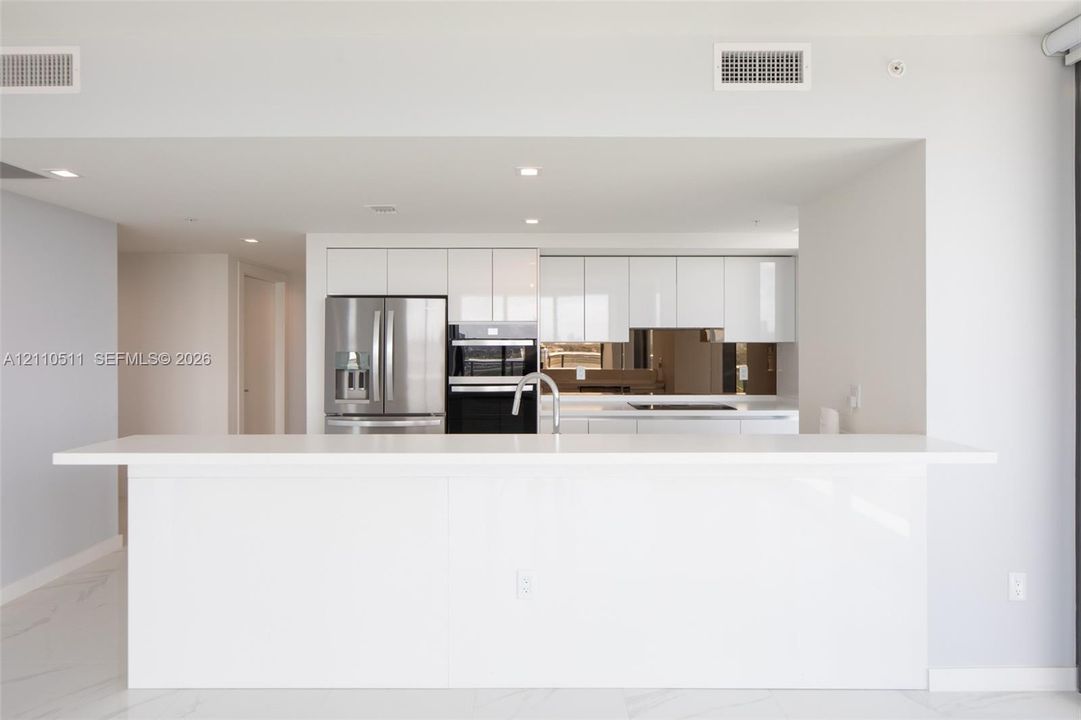 Modern kitchen featuring high-gloss white cabinetry, expansive stone-finish peninsula, stainless steel french-door refrigerator, integrated double oven, and mirrored tile backsplash