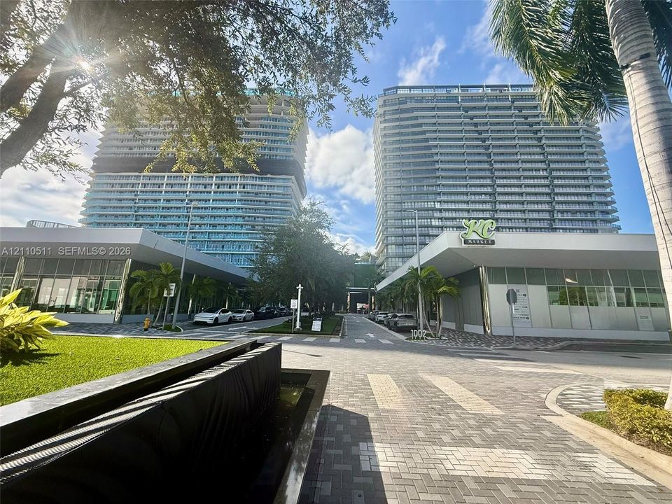 Modern high-rise towers featuring floor-to-ceiling glass facades, covered ground-level commercial spaces, paved stone driveways, and landscaped green areas