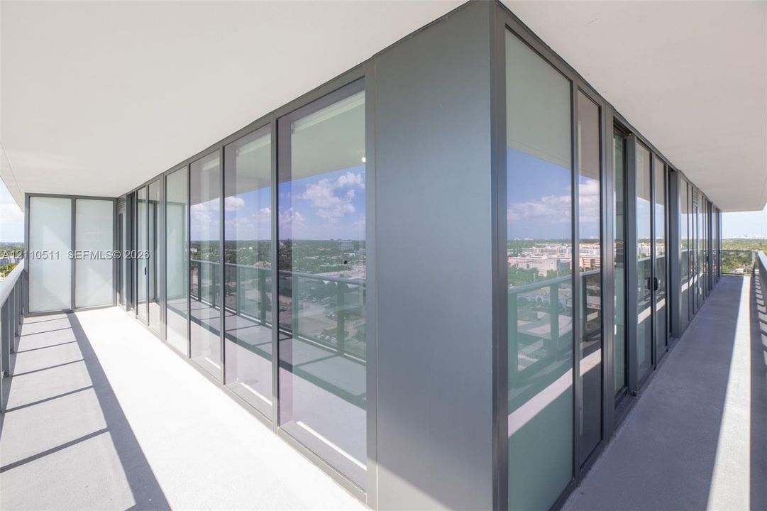 Wrap-around exterior balcony featuring floor-to-ceiling glass panels, dark metal framing, concrete flooring, and minimalist railing