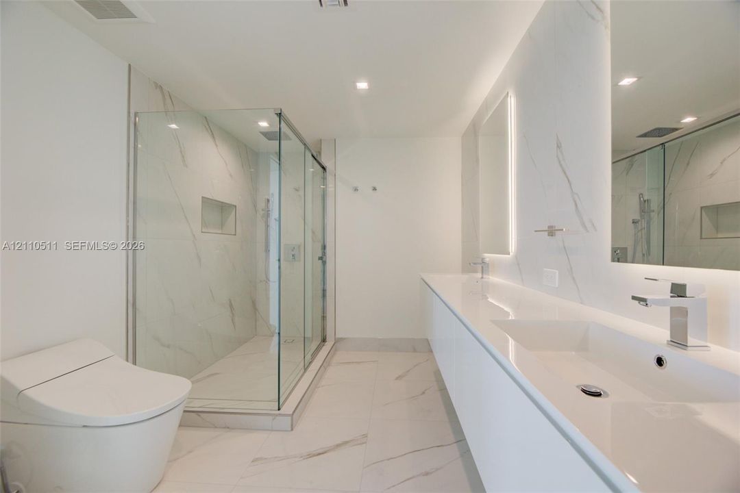Bathroom featuring a dual-sink vanity with a solid-surface countertop, walk-in glass-enclosed shower with wall-mounted plumbing fixtures, recessed shower niche, and large-format porcelain tile flooring