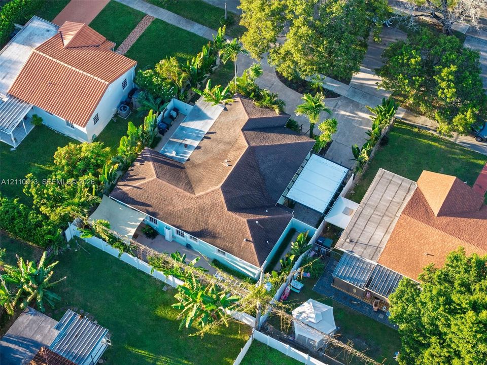 Aerial perspective of a property featuring an asphalt shingle roof, a concrete driveway, a screened-in swimming pool enclosure, and lush tropical landscaping