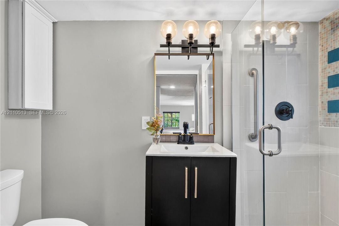 Bathroom featuring a vanity with dark cabinetry and a light-toned countertop, three-light fixture, gold-framed mirror, and a glass-enclosed shower with tiled surround