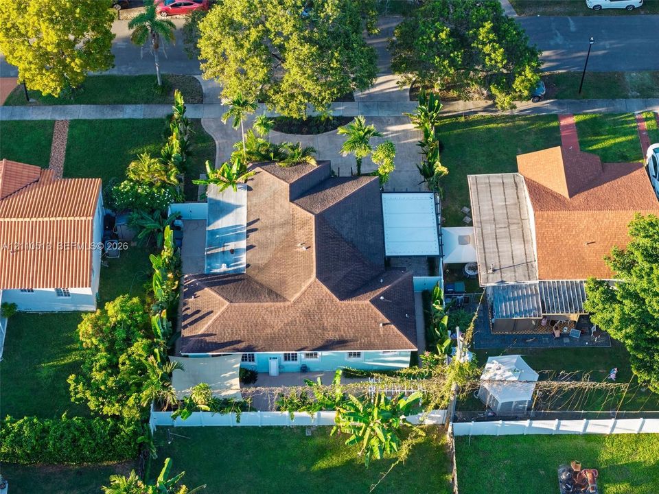 Aerial perspective featuring a multi-hipped roofline, paved driveway, covered patio area, white vinyl perimeter fencing, and mature landscaping