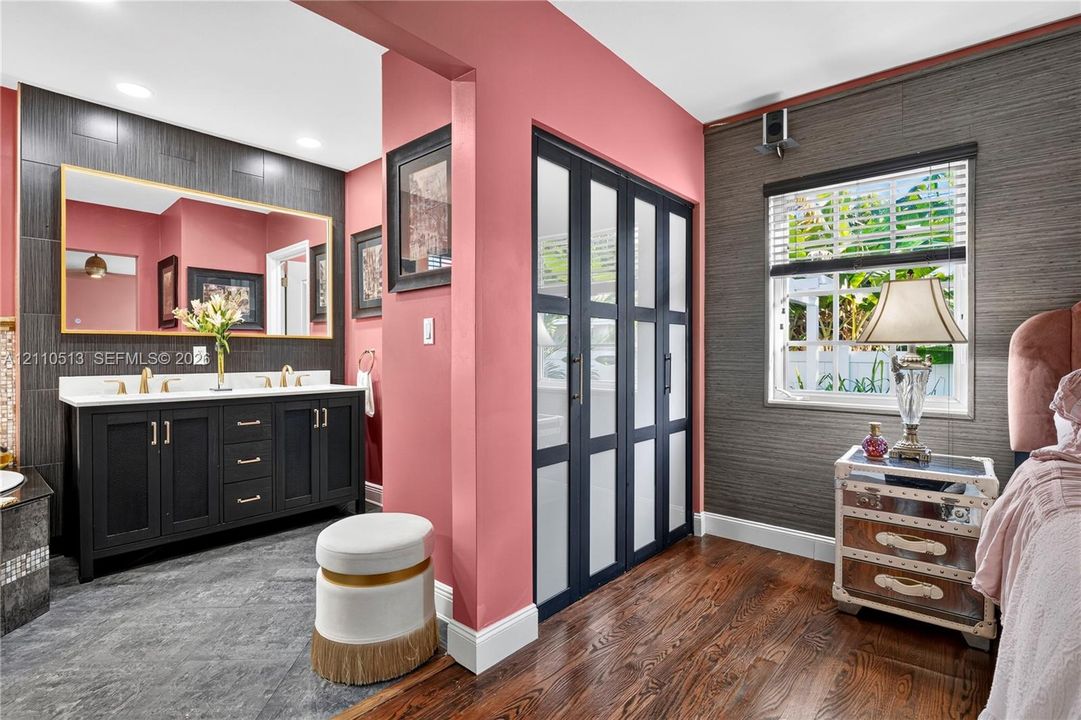 Double vanity featuring a white countertop and gold-toned hardware, large rectangular wall mirror, wood-finish flooring, dark-toned textured wall treatment, and floor-to-ceiling glass-paneled closet doors