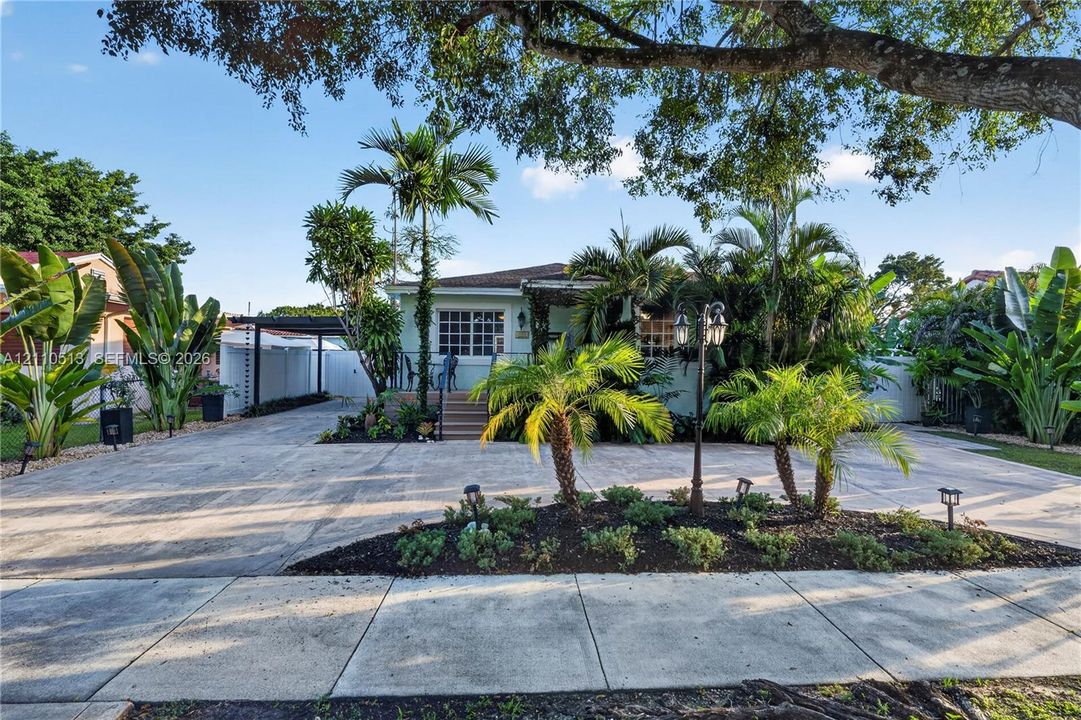 Exterior facade featuring a covered entryway, paved driveway, carport, and landscape elements including palm trees and mulched garden beds