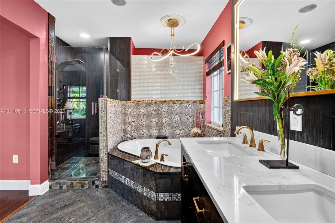Bathroom featuring a dual-sink vanity with white stone-finish countertops, gold-toned fixtures, a corner jetted soaking tub with mosaic tile surround, and an enclosed shower with dark tile walls