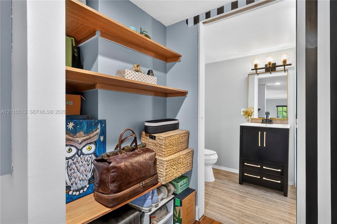 Built-in wall shelving, modern black vanity with gold-tone hardware, rectangular mirror with integrated vanity lighting, and wood-finish flooring