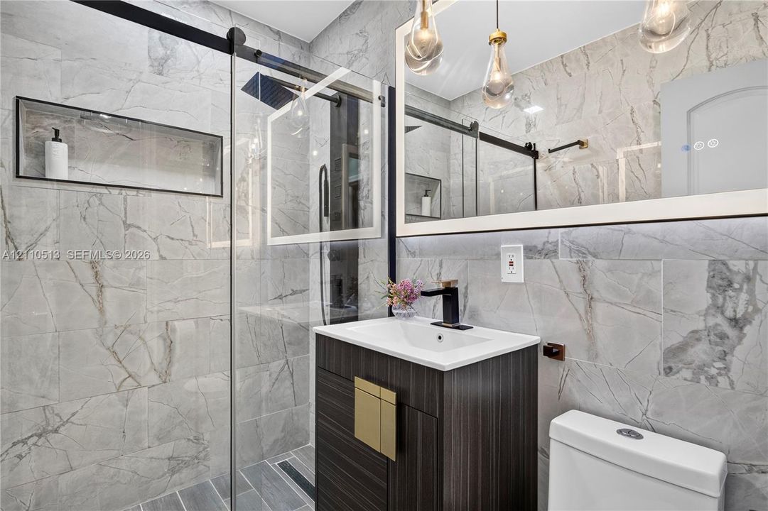 Bathroom featuring full-height large-format stone tile, a glass-enclosed shower with a built-in recessed wall niche, a dark wood-finish vanity with a rectangular sink, matte black fixtures, and a framed mirror with pendant lighting