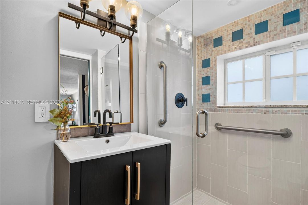 Dark cabinetry vanity with a white countertop, metallic hardware, rectangular framed mirror with integrated light fixture, and glass-enclosed shower with tiled accents