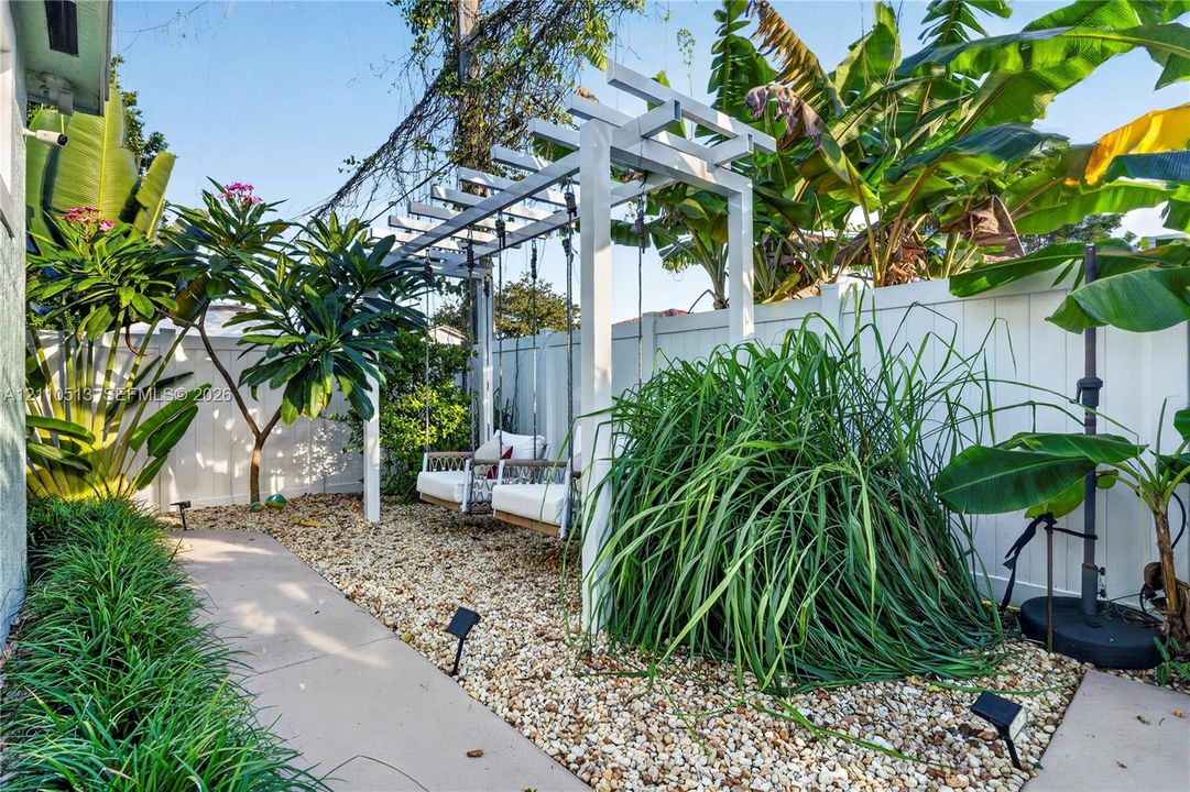 Custom white wooden pergola, gravel landscaping, concrete pathway, mature tropical vegetation, and perimeter vinyl fencing