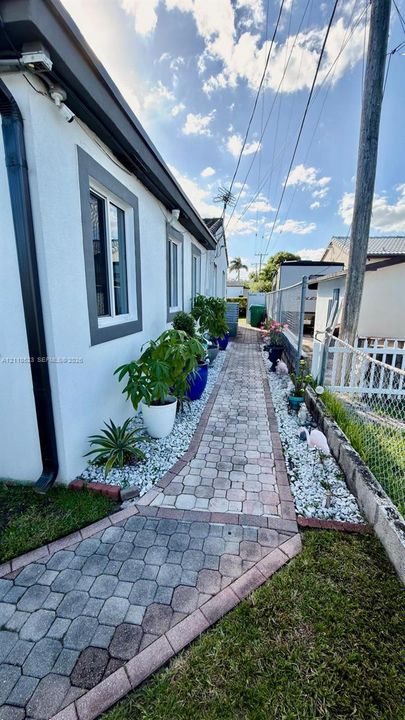Side exterior featuring a paver walkway bordered by white stone landscaping, white stucco siding with grey trim, and metal chain-link fencing
