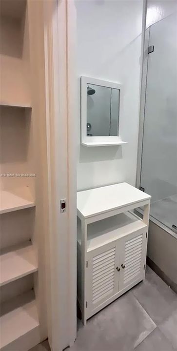 White vanity with louvered cabinet doors, open shelving, and integrated mirror, accompanied by built-in wall shelving and a glass-enclosed shower
