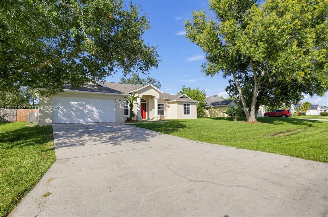 Single-story exterior featuring a stucco facade, concrete driveway, two-car garage, covered entryway with columns, and established shade trees
