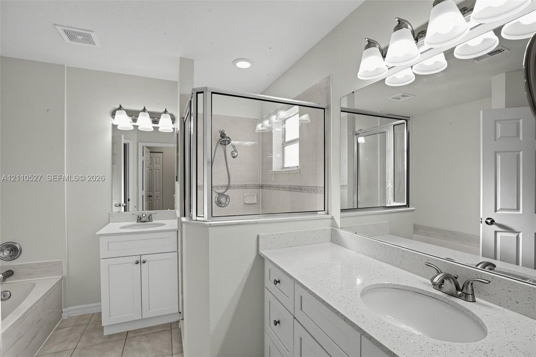 Bathroom featuring dual white shaker-style cabinetry, light-toned stone-finish countertops, an enclosed glass-paneled shower with tiled surround, and large-scale vanity mirrors with multi-bulb light fixtures