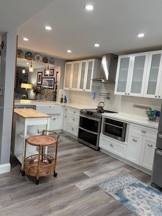 Kitchen featuring white cabinetry with glass-front uppers, stone-finish countertops, apron-front sink, stainless steel range hood, and wood-finish flooring