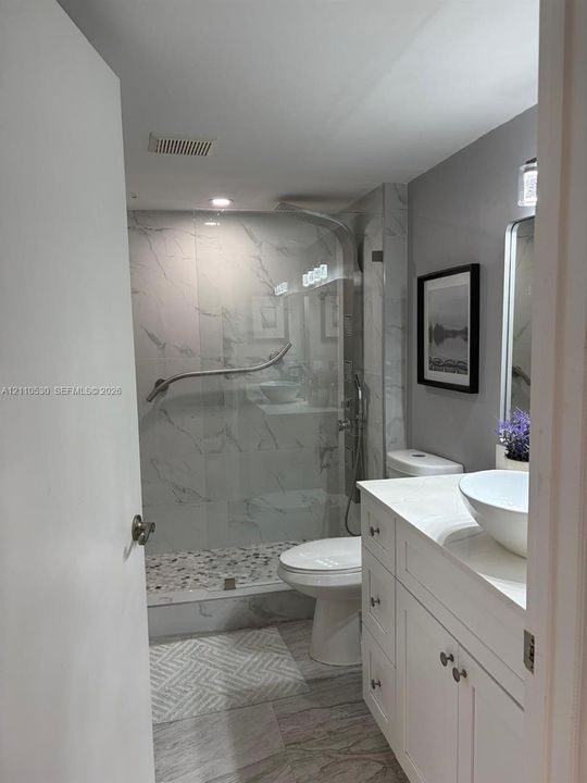 Bathroom featuring a white vanity cabinet with an integrated white vessel sink, walk-in shower with stone-tile flooring and glass enclosure, and stone-finish wall tile