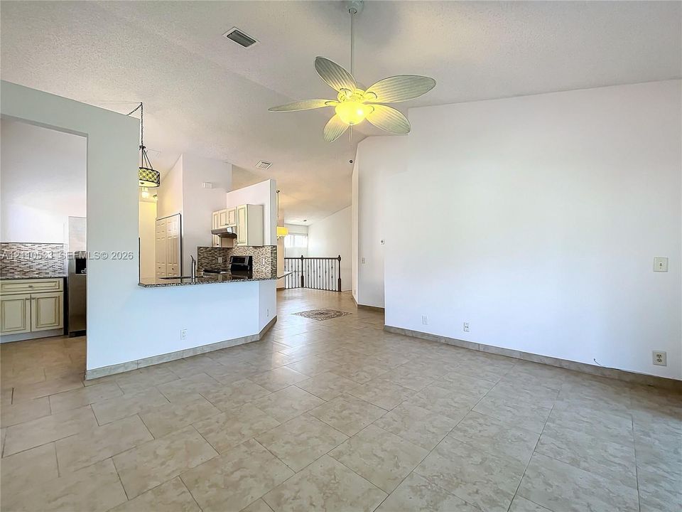 Open-concept floor plan featuring tile flooring, vaulted ceilings, a ceiling fan with integrated lighting, a kitchen breakfast bar, and iron stair railing