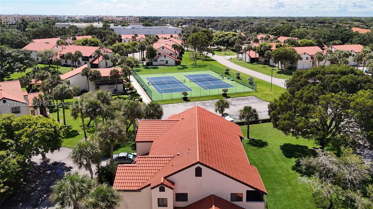 Aerial perspective featuring red tile roofing, expansive manicured grounds with mature trees, and dual fenced tennis courts