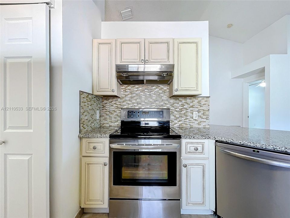 Kitchen featuring cream-colored cabinetry with brushed hardware, stone-patterned countertops, a stainless steel range with a built-in exhaust hood, and a decorative tile backsplash