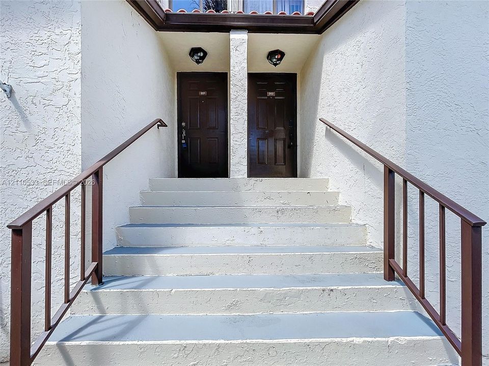 Exterior entrance featuring concrete steps, dual solid wood-finish entry doors, metal handrails, and surface-mounted light fixtures