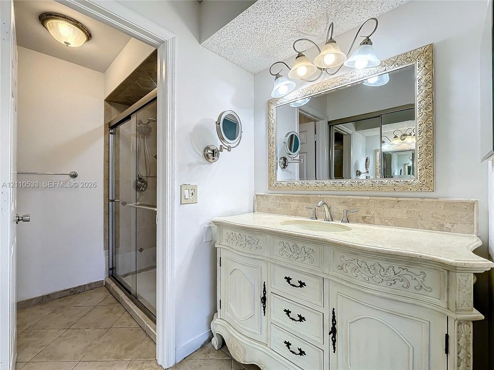 Vanity featuring ornate cabinetry, natural stone countertop, and integrated sink with widespread faucet, accompanied by a shower enclosure with tile surround and a wall-mounted magnifying mirror