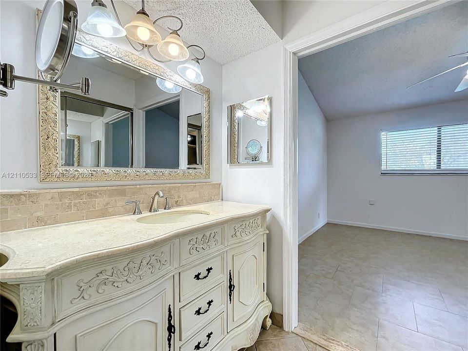 Vanity featuring ornate cabinetry, light-toned stone countertop with an integrated basin, tile backsplash, and decorative-frame wall mirror with multi-bulb light fixture
