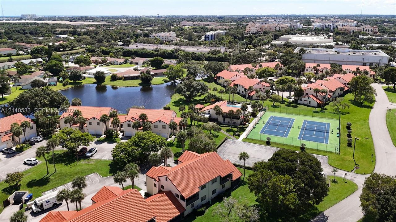 Aerial perspective of the community featuring dual tennis courts, a central pond, multi-story buildings with terracotta-style roof tiles, and mature landscaping