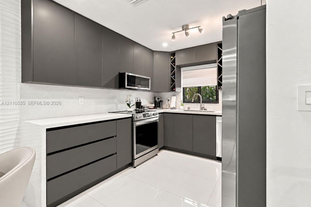 Modern kitchen featuring charcoal slab-panel cabinetry, white solid-surface countertops, integrated wine storage, stainless steel appliances, and large-format porcelain tile flooring