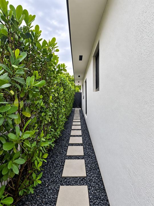 Exterior side pathway featuring a white stucco facade, concrete paver stones, dark stone ground cover, and a manicured hedge border