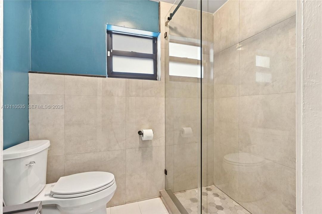Walk-in shower with a glass enclosure, large-format neutral wall tile, hexagonal floor tile, and window with dark trim