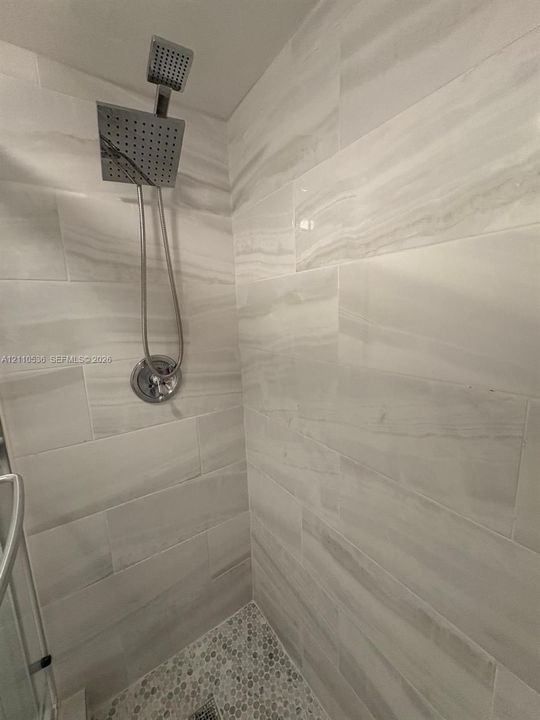 Shower stall featuring large-format stone-patterned wall tile, pebble-texture floor tile, and a polished chrome square rainfall showerhead with a handheld sprayer