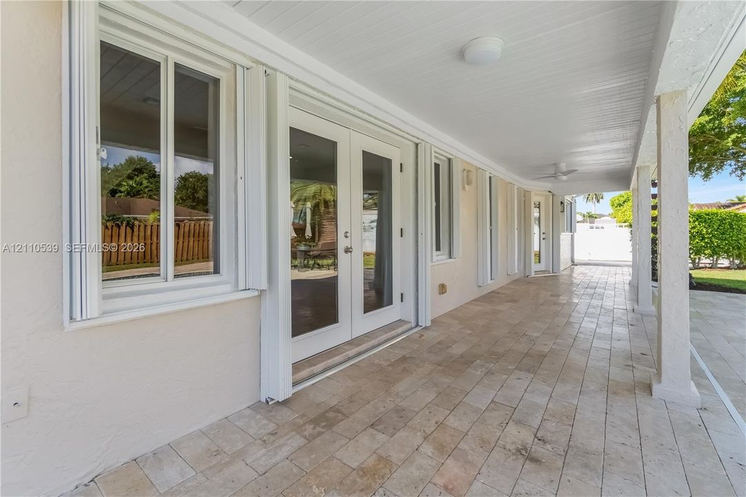 Covered patio featuring stone-tile flooring, glass-paned double doors, white-trimmed windows, and ceiling-mounted light fixtures