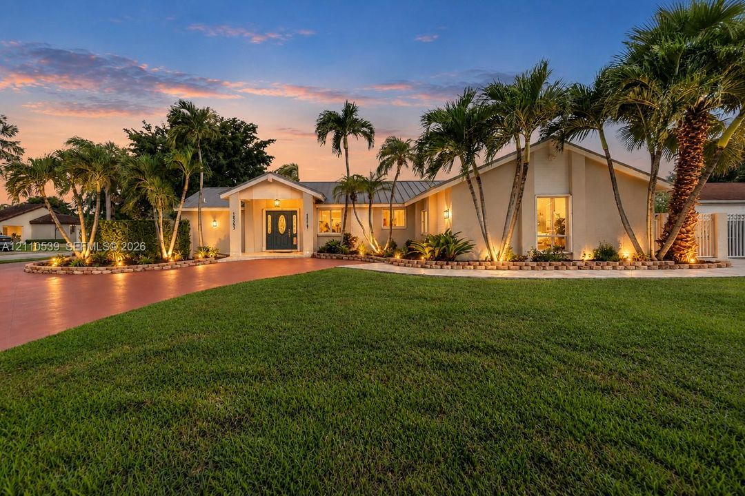 Stucco exterior featuring a covered entryway, metal roofing, brick-edged landscaping, mature palm trees, and a paved driveway