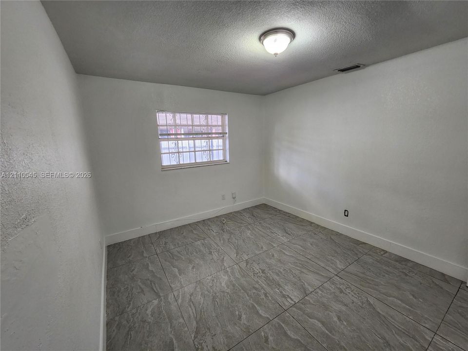 Interior space featuring large-format stone-patterned tile flooring, crisp white wall finishes, a single-hung window with security bars, and a central ceiling-mounted flush light fixture