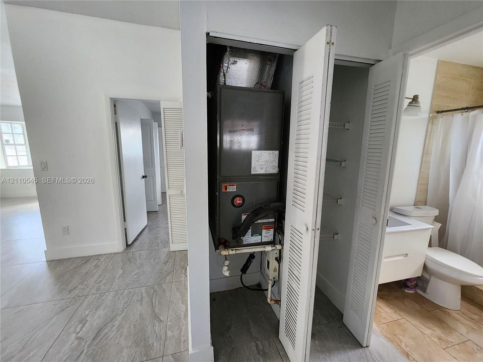 Utility closet featuring a central HVAC unit and integrated shelving, accompanied by floor-to-ceiling tile in the sanitation space, a wall-mounted vanity, and ceramic tile flooring