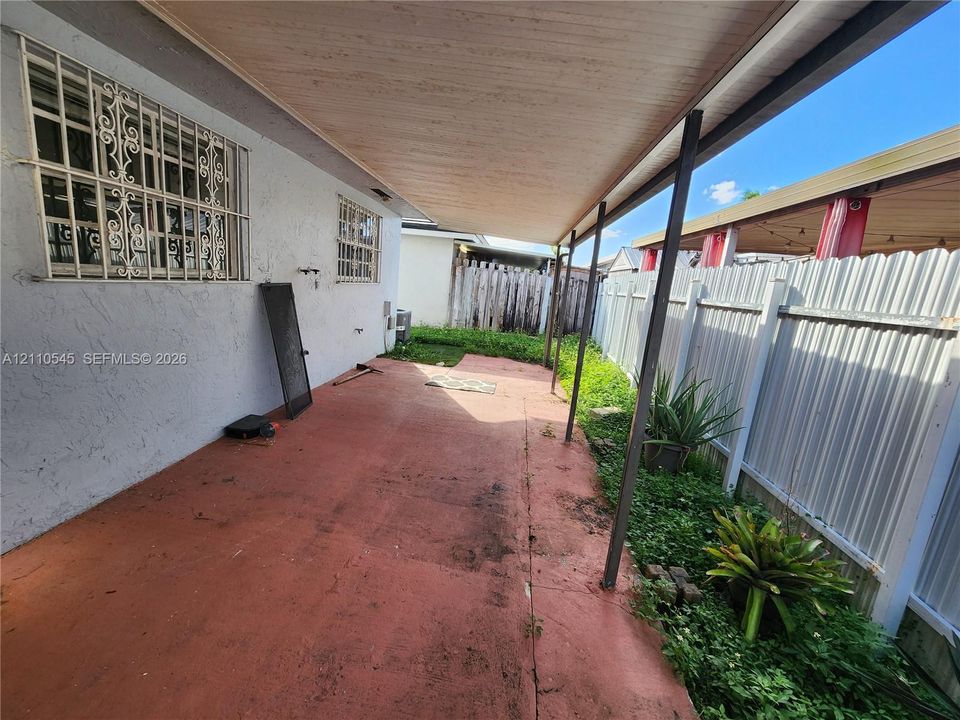 Covered patio featuring concrete flooring, stucco exterior walls, metal security window bars, and corrugated metal perimeter fencing