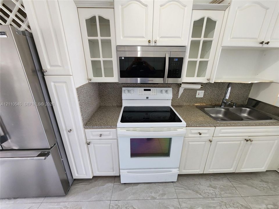 White cabinetry featuring glass-paneled uppers, stone-composite countertops, stainless steel refrigerator, over-the-range microwave, and ceramic tile flooring