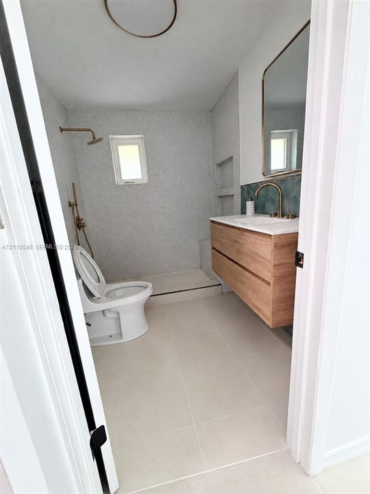 Floating wood-finish vanity with a white countertop, wall-mounted brass faucet, arched vanity mirror, and tiled shower surround with brass fixtures