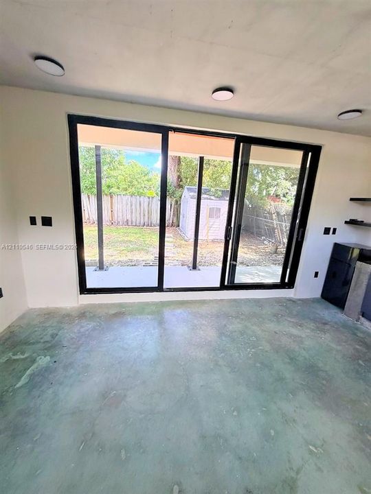 Large floor-to-ceiling black-framed sliding glass doors opening to a concrete patio, polished concrete flooring, and flush-mount ceiling lighting