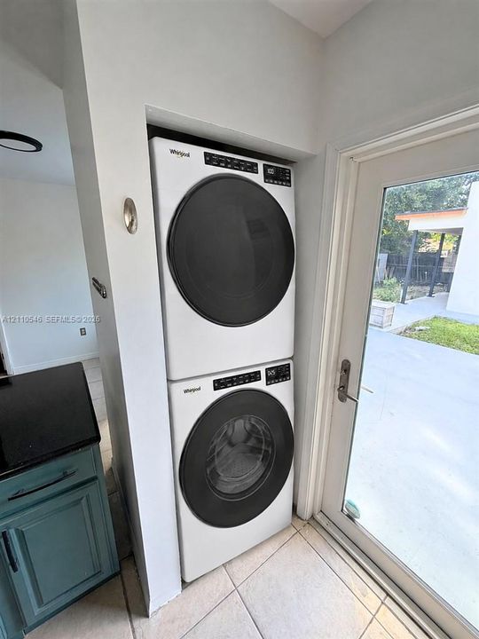 Stacked high-efficiency front-load washer and dryer unit with digital control panels, tile flooring, and glass-paneled exterior door