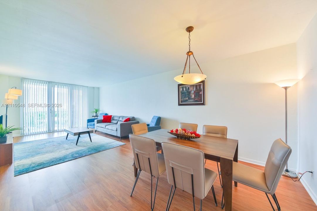 Open-concept area featuring wood-finish flooring, a suspended metal-frame pendant light fixture, and floor-to-ceiling sliding glass doors with vertical blinds