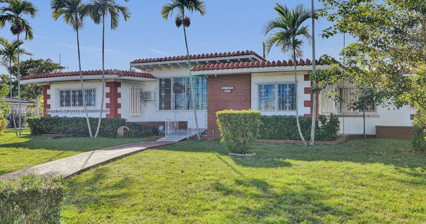 Single-story residence featuring a tile roof, brick masonry accents, white stucco exterior, and a paved walkway leading to the entryway