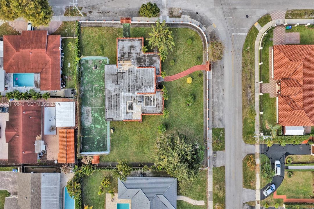 Aerial perspective of a corner lot property featuring a private tennis court, expansive lawn, perimeter masonry wall, and paved walkway