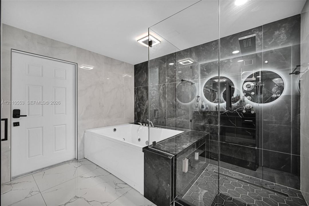 Jetted soaking tub, walk-in shower with glass enclosure, large-format tile flooring, polished stone wall surfacing, and dual illuminated circular mirrors