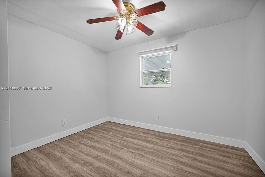 Interior room featuring wood-finish flooring, neutral wall tones, a ceiling fan with multi-light fixture, and a single window with blinds